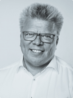 Foto von Dirk Zieschang, Schwarz-Weiß-Foto eines Mannes mit kurzen Haaren und Brille, lächelnd, in einem weißen Hemd.