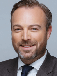 Foto von Florian Stumm, Porträt eines Mannes mit Bart, in Anzug und Krawatte, lächelnd, vor einem neutralen Hintergrund.