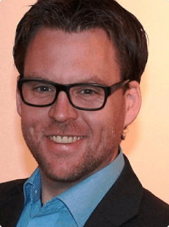 Foto von Sven Wissman, Porträt eines Mannes mit kurzen Haaren und Brille, der breit lächelt, in einem Jackett, vor einem neutralen Hintergrund.