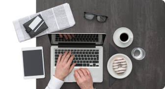 Draufsicht auf einen Arbeitsplatz mit Händen, die auf einem Laptop tippen. Links liegen eine Zeitung und eine Brille, auf dem Schreibtisch liegt ein Tablet, darauf eine schwarze Lederbrieftasche. Rechts liegen eine Tasse Kaffee und ein Teller mit einem Donut mit weißer Glasur und Schokoladenüberzug.